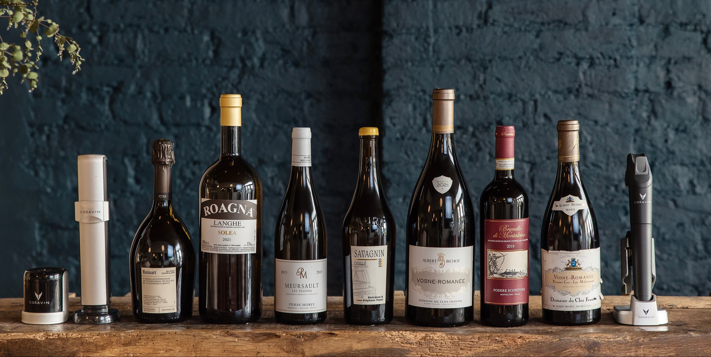 A curated selection of fine wines, including Meursault, Roagna Langhe, and Vosne-Romanée, displayed with Coravin wine preservation systems on a rustic wooden table against a dark brick wall.