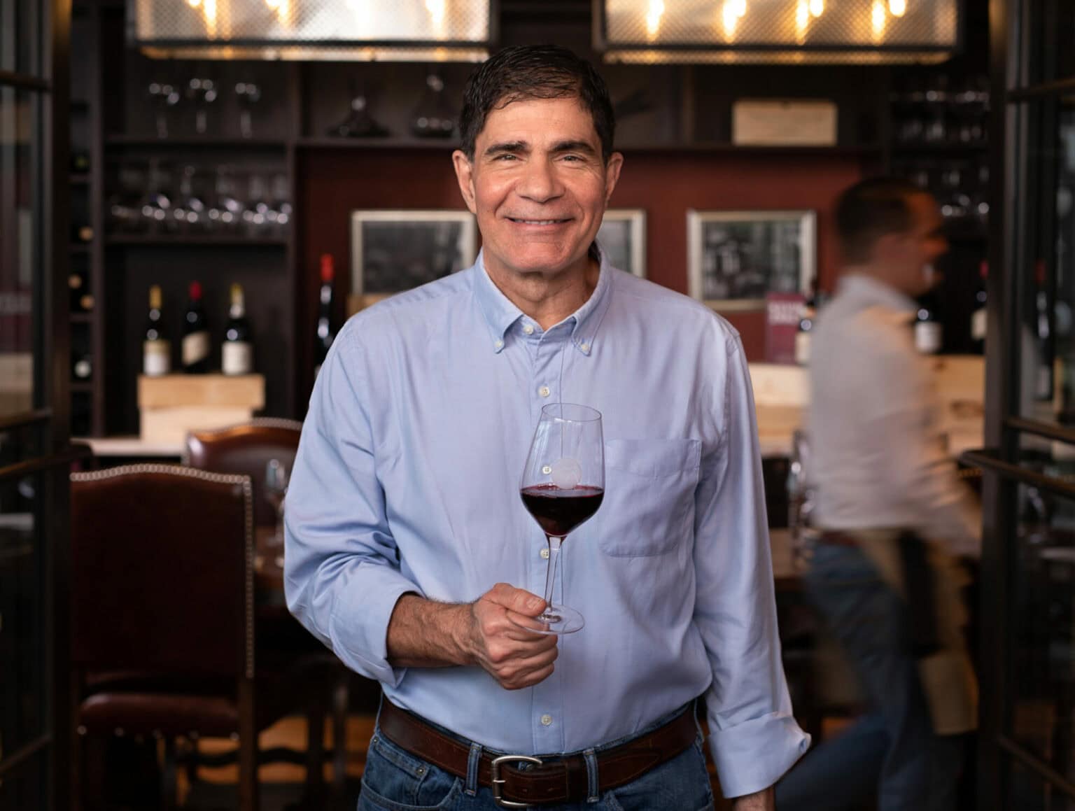 A man in a light blue shirt smiles while holding a glass of red wine in a restaurant setting with wine bottles in the background.