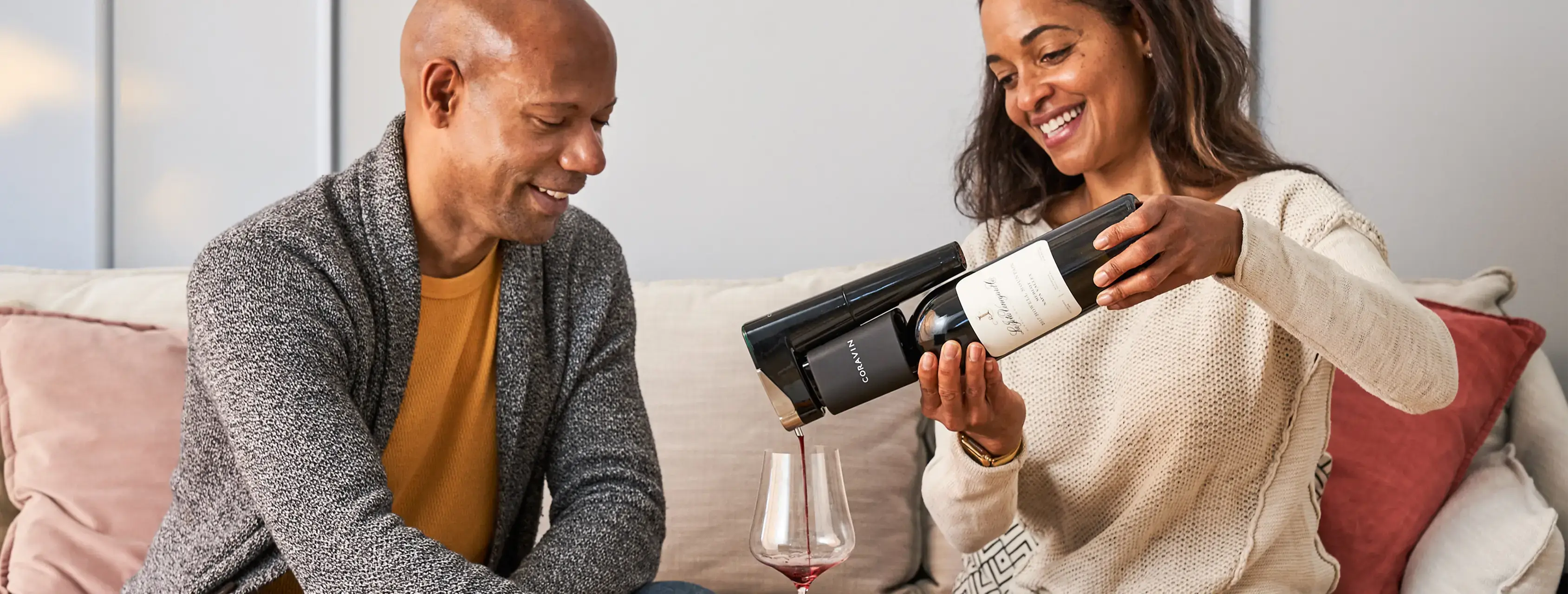 A couple enjoys pouring wine from a bottle using a Coravin wine preservation system while relaxing on a couch.