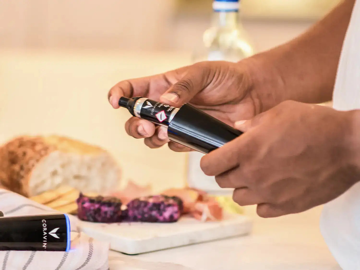 A person holding a Coravin wine preservation tool with bread and a charcuterie board in the background.