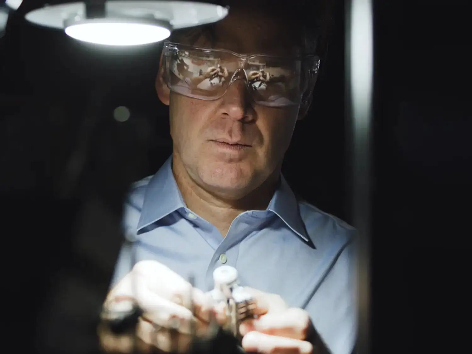 A person wearing safety glasses focuses on assembling small mechanical components under a bright lamp.