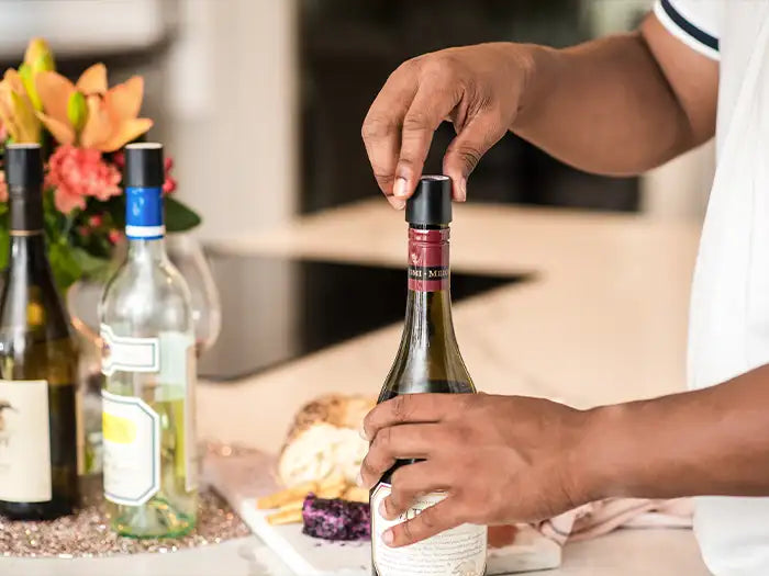 A person removes the foil from a wine bottle, with various wines and a platter of cheese and snacks nearby.