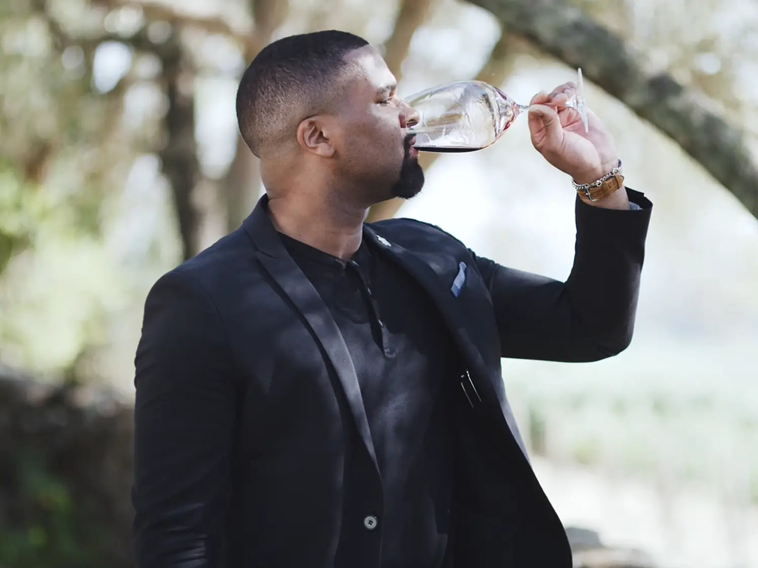 Man in a black blazer sipping red wine from a glass outdoors, surrounded by trees and greenery.