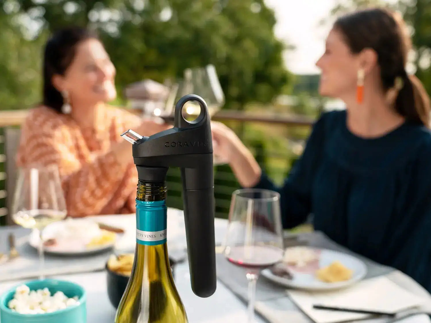 A Coravin wine aerator is positioned on a bottle, with two women chatting and enjoying food and wine in the background.