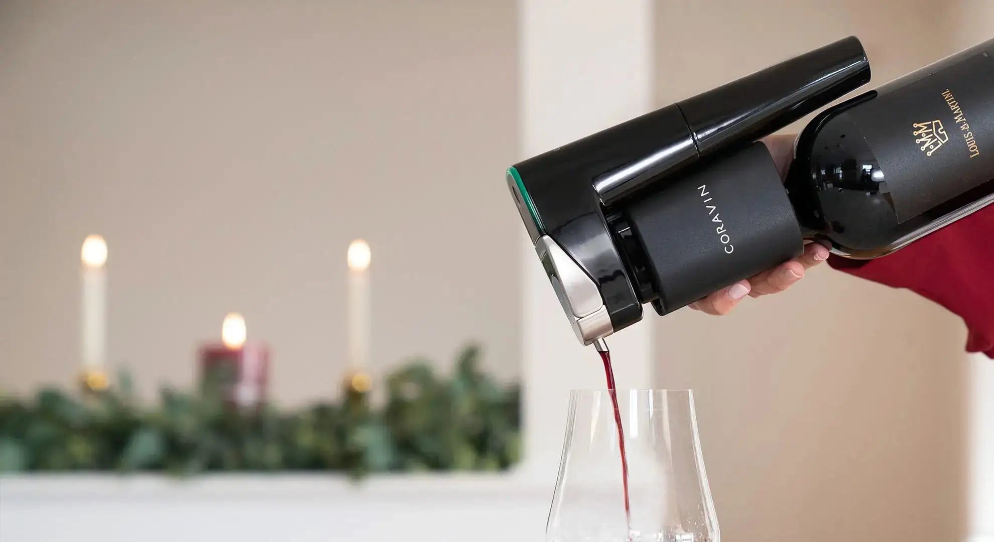 A Coravin wine preserver pours red wine into a glass, with candles and greenery in the background.