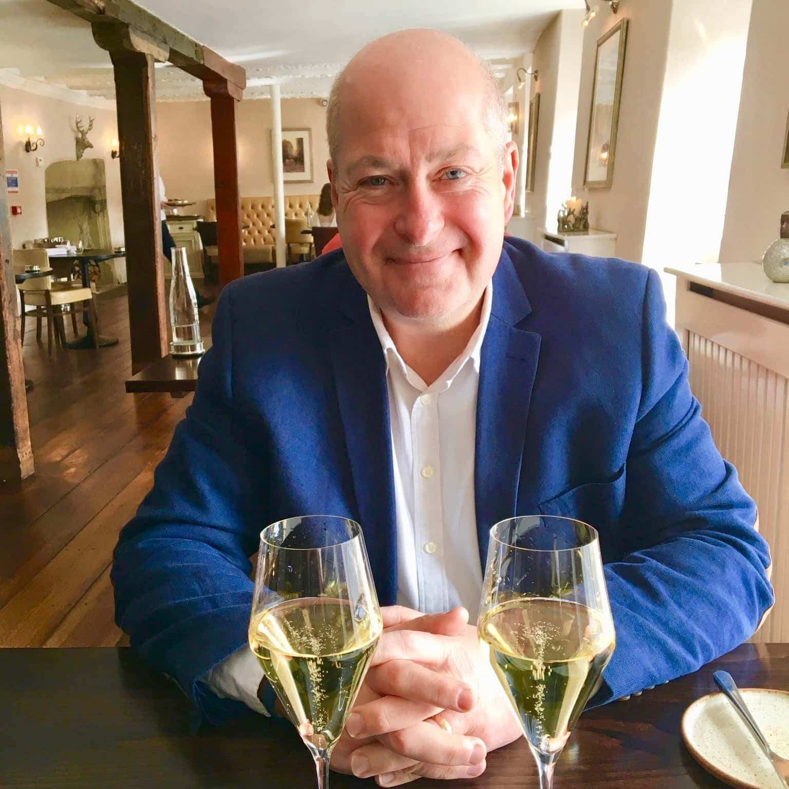 Man smiling warmly at a table with two glasses of sparkling wine, set in a stylish dining room.