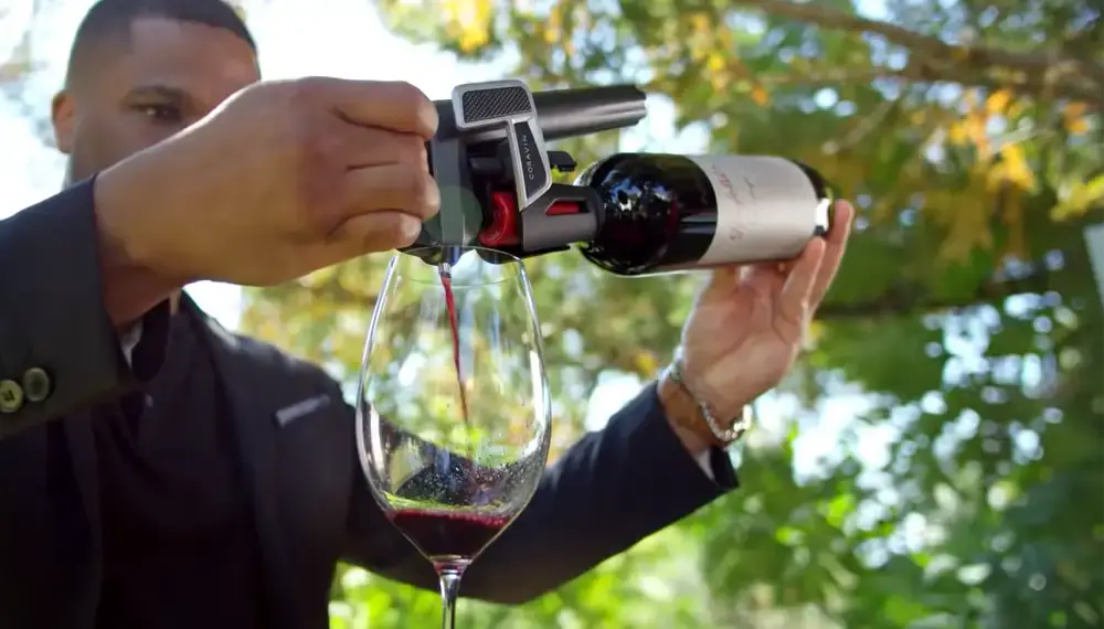 A person pours red wine from a bottle into a glass using a wine pouring tool, surrounded by greenery.