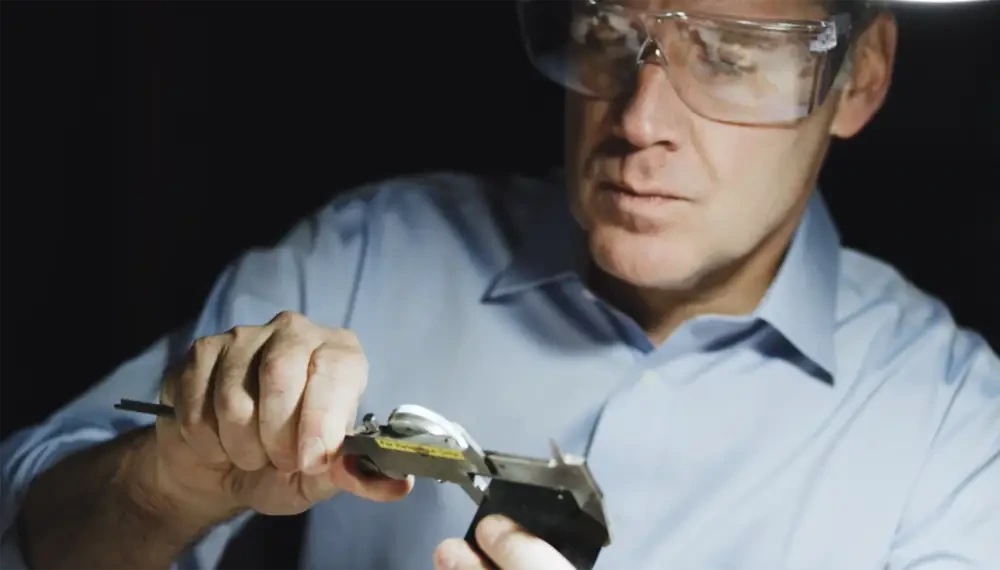 Man wearing safety glasses meticulously measures an object using a caliper in a dimly lit environment.
