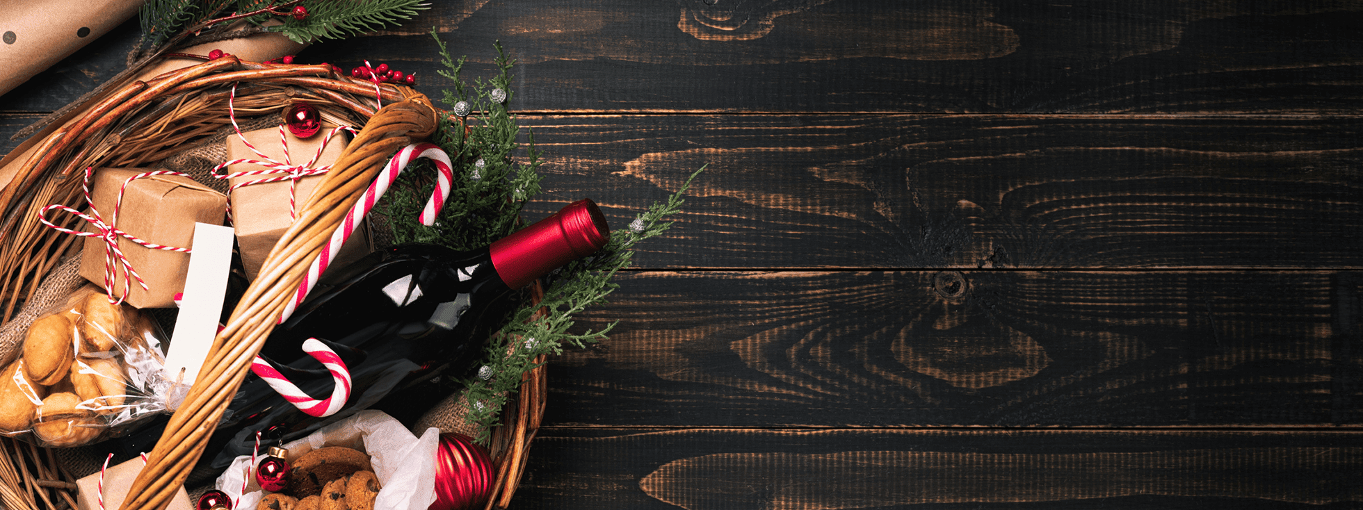 Festive holiday gift basket with a bottle of red wine, candy canes, cookies, and wrapped presents on a rustic wooden background.