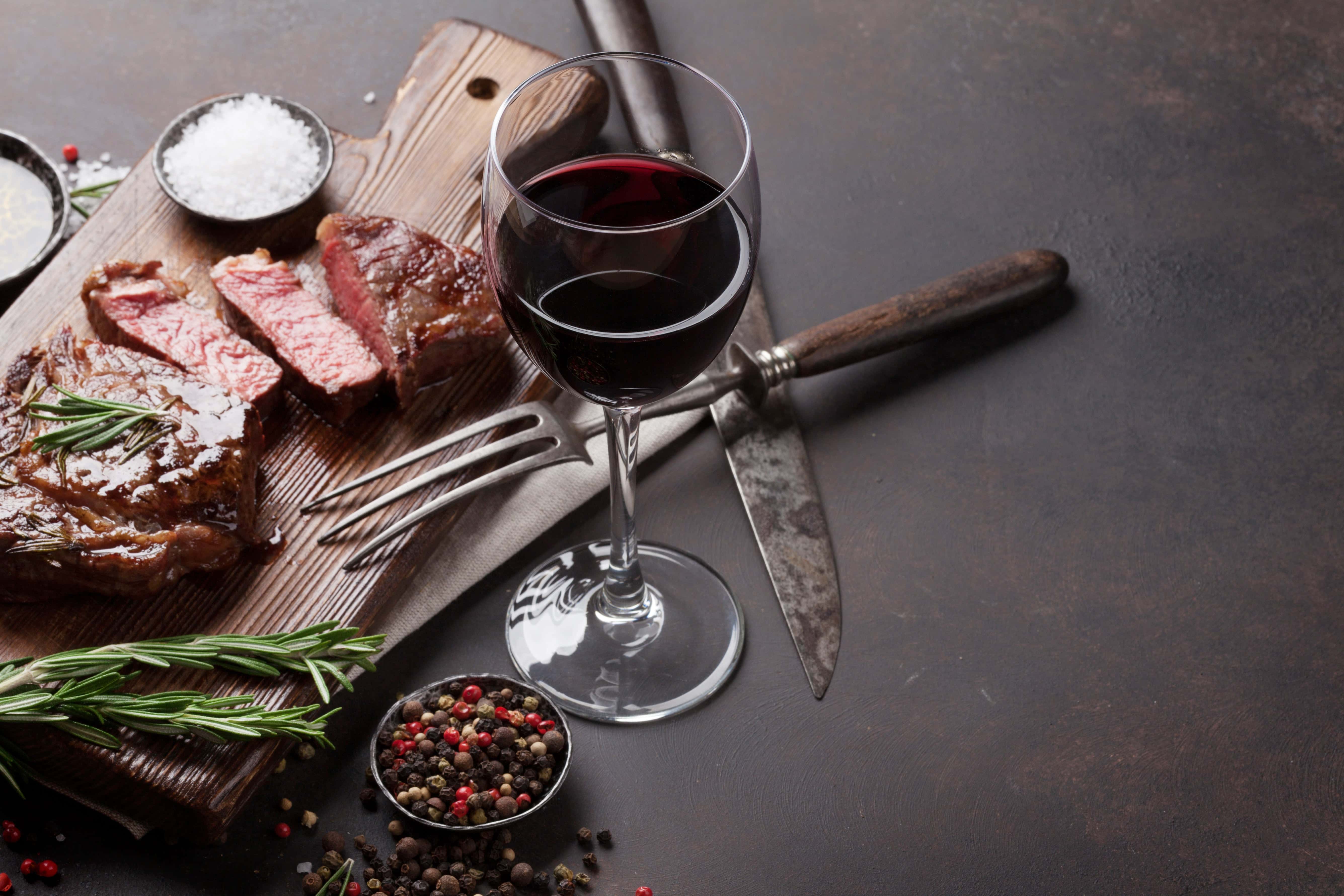 Glass of red wine served beside a sliced grilled steak on a wooden board, garnished with rosemary and accompanied by salt, peppercorns, and vintage cutlery on a dark rustic background.