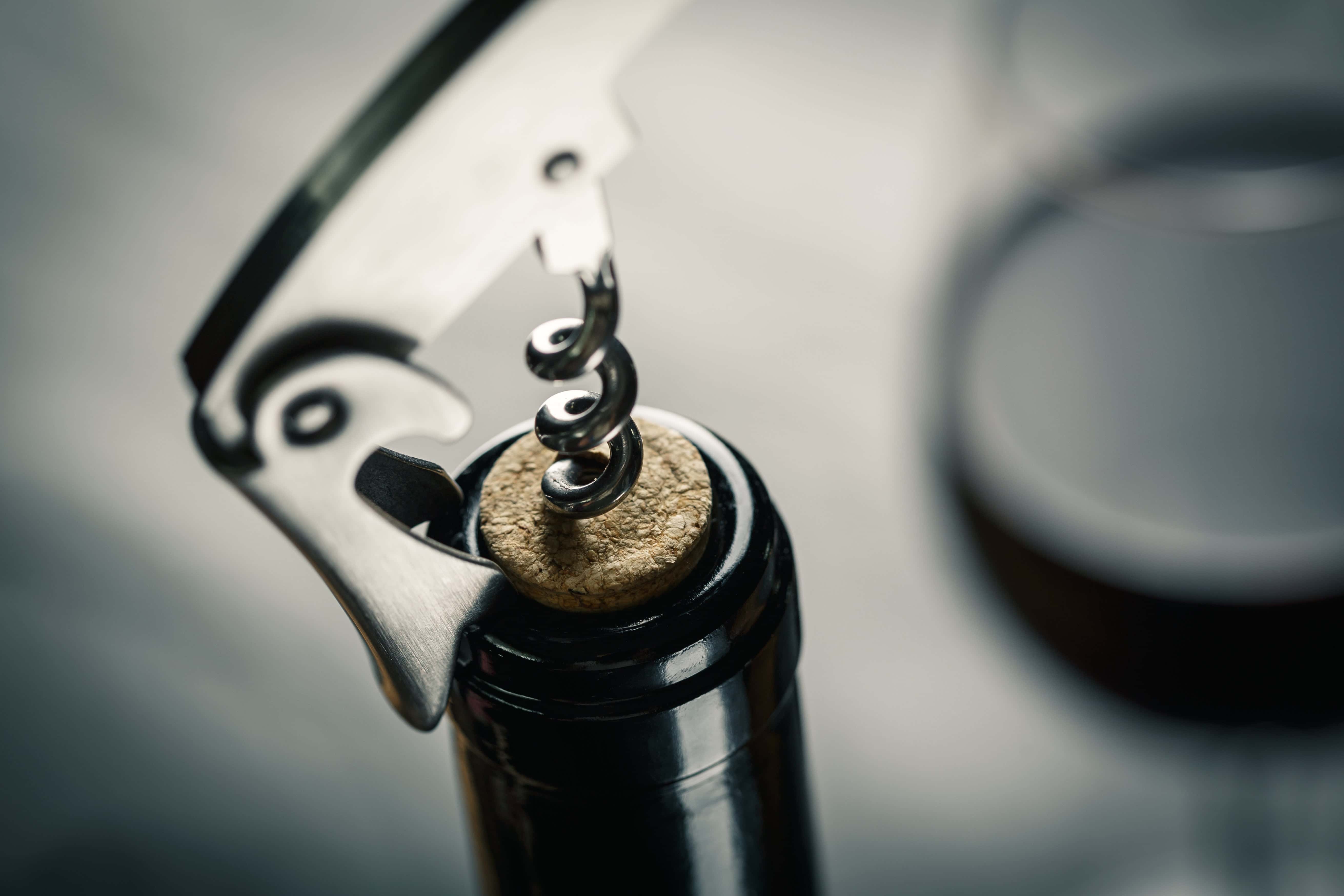 Close-up of a corkscrew being inserted into a wine bottle cork, with a glass of red wine blurred in the background.