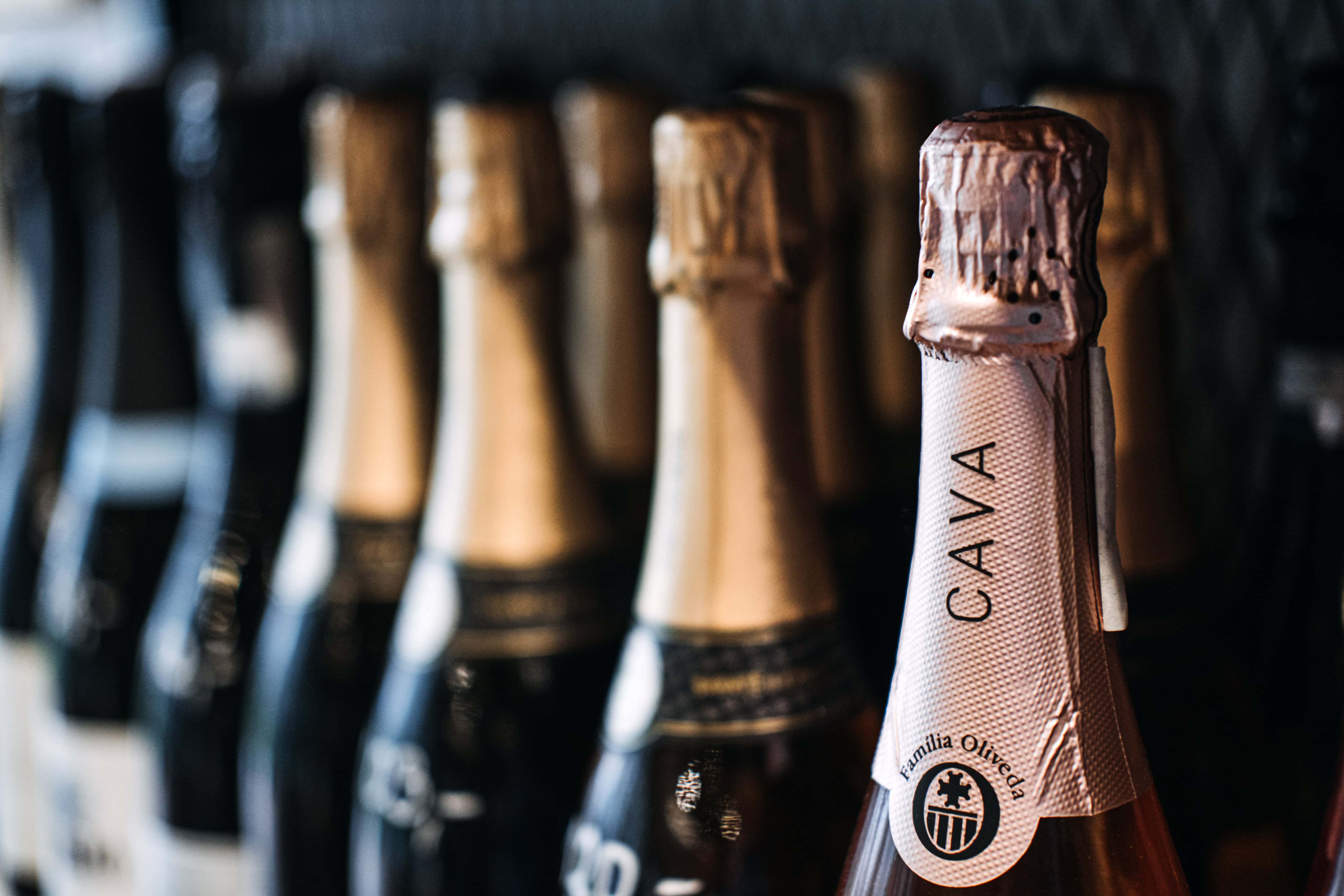 Close-up of a row of sparkling wine bottles, with one labeled 'Cava' in focus, featuring foil-wrapped tops and a dimly lit background.