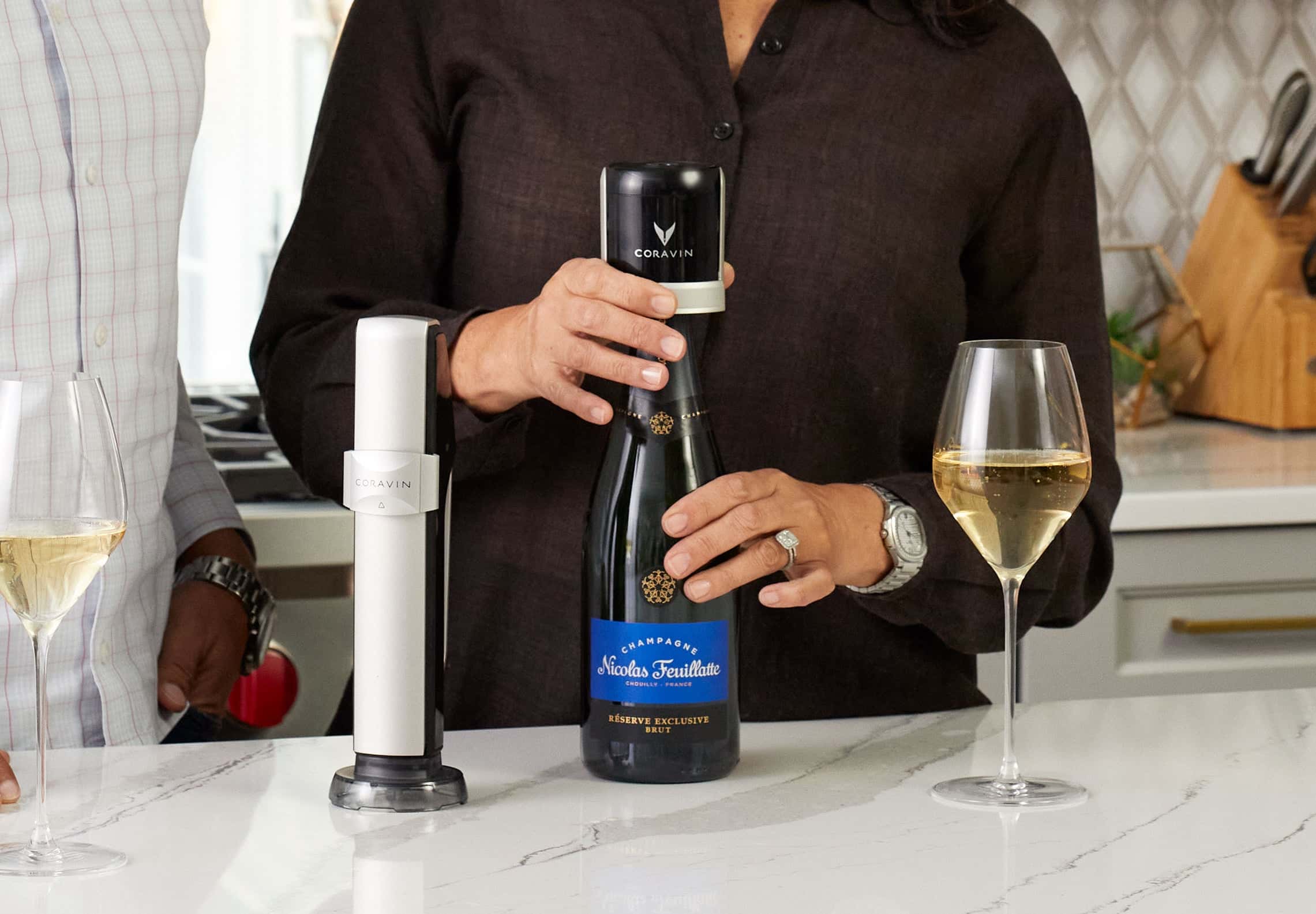 Hands sealing a bottle of Nicolas Feuillatte Champagne with the Coravin Sparkling Wine Preservation System, on a modern kitchen counter with full glasses of sparkling wine.