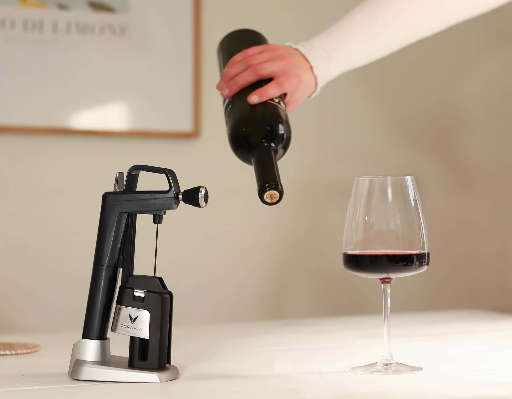 Coravin Timeless Six wine preservation system in Piano Black next to a glass of red wine, with a hand holding a cork-sealed bottle on a neutral indoor background.