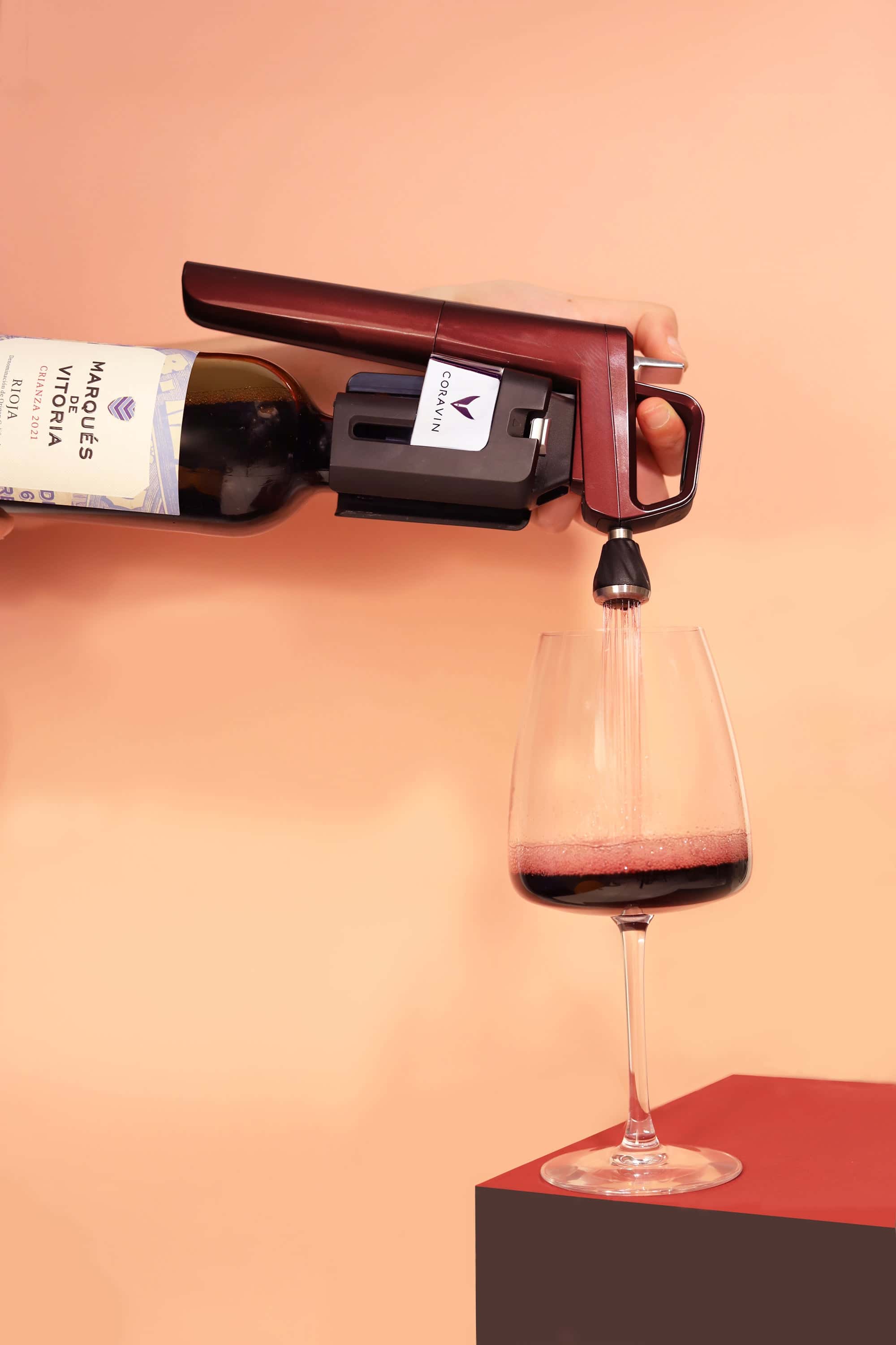 Coravin wine preservation system pouring red wine from a Marqués de Vitoria Rioja bottle into a glass against a peach-colored background.