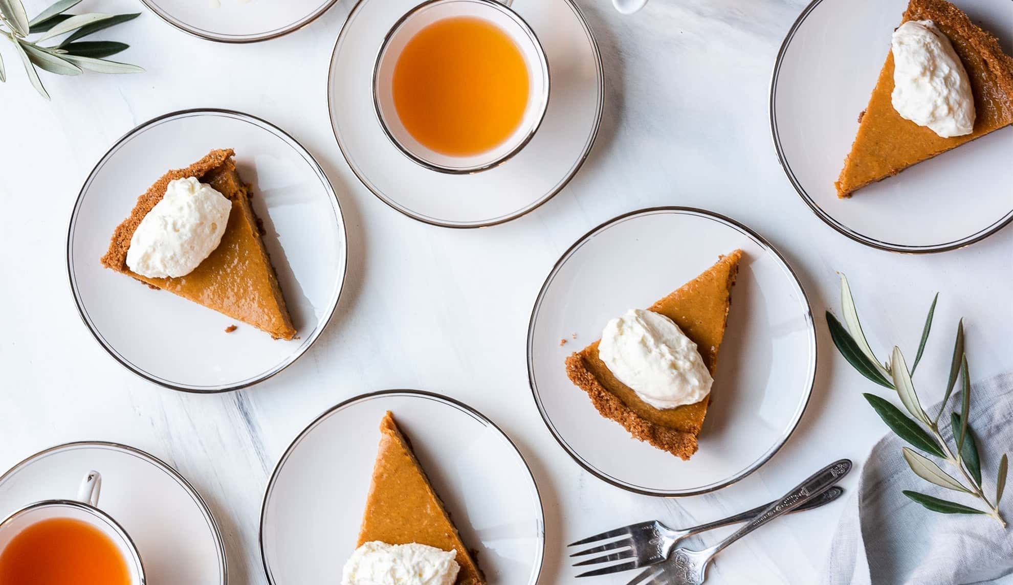 Slices of pumpkin pie with whipped cream served on white plates, paired with cups of tea on a white marble table setting.