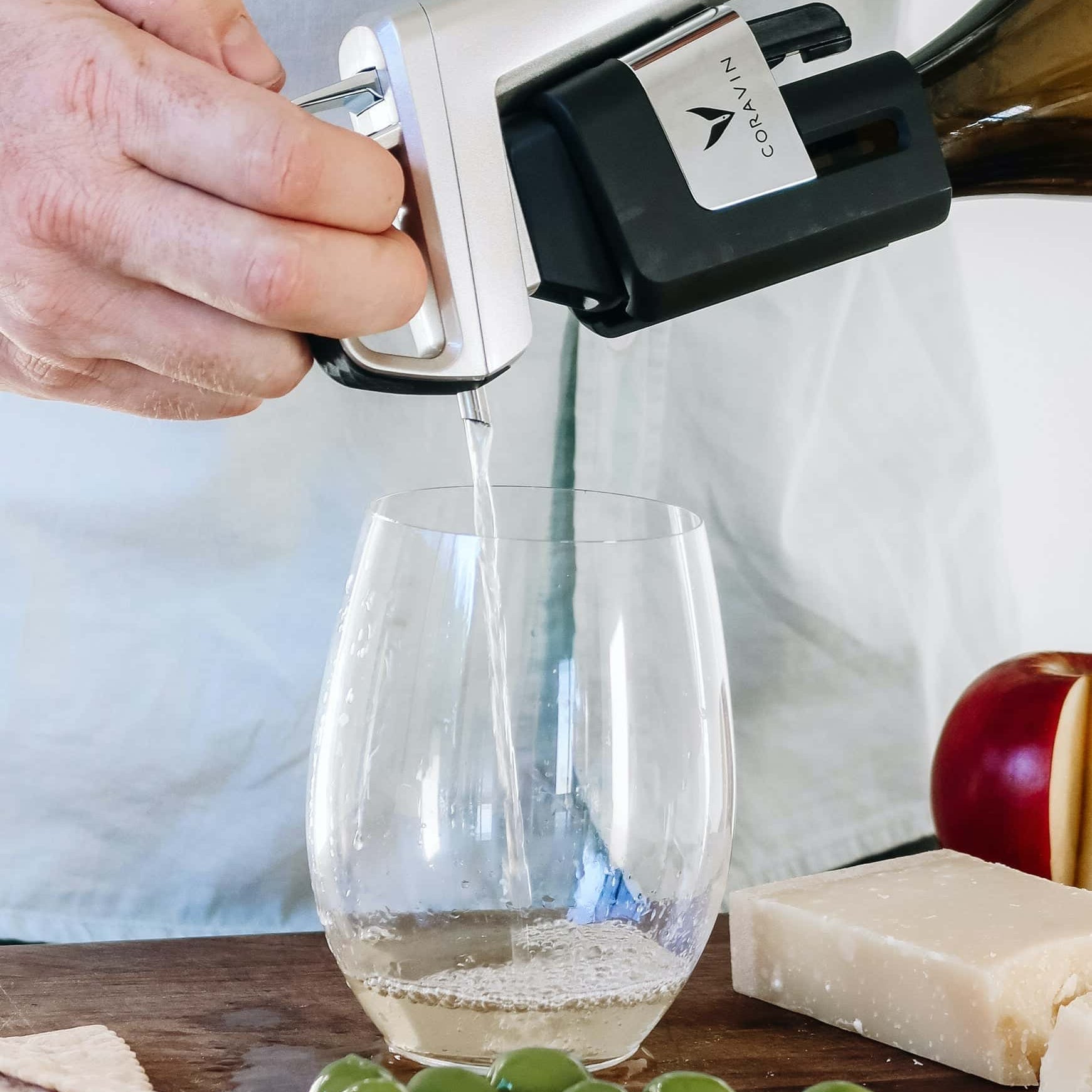 Close-up of a hand pouring white wine using a Coravin wine preservation system into a stemless glass, with green olives, crackers, a wedge of cheese, and a sliced apple on a wooden surface.