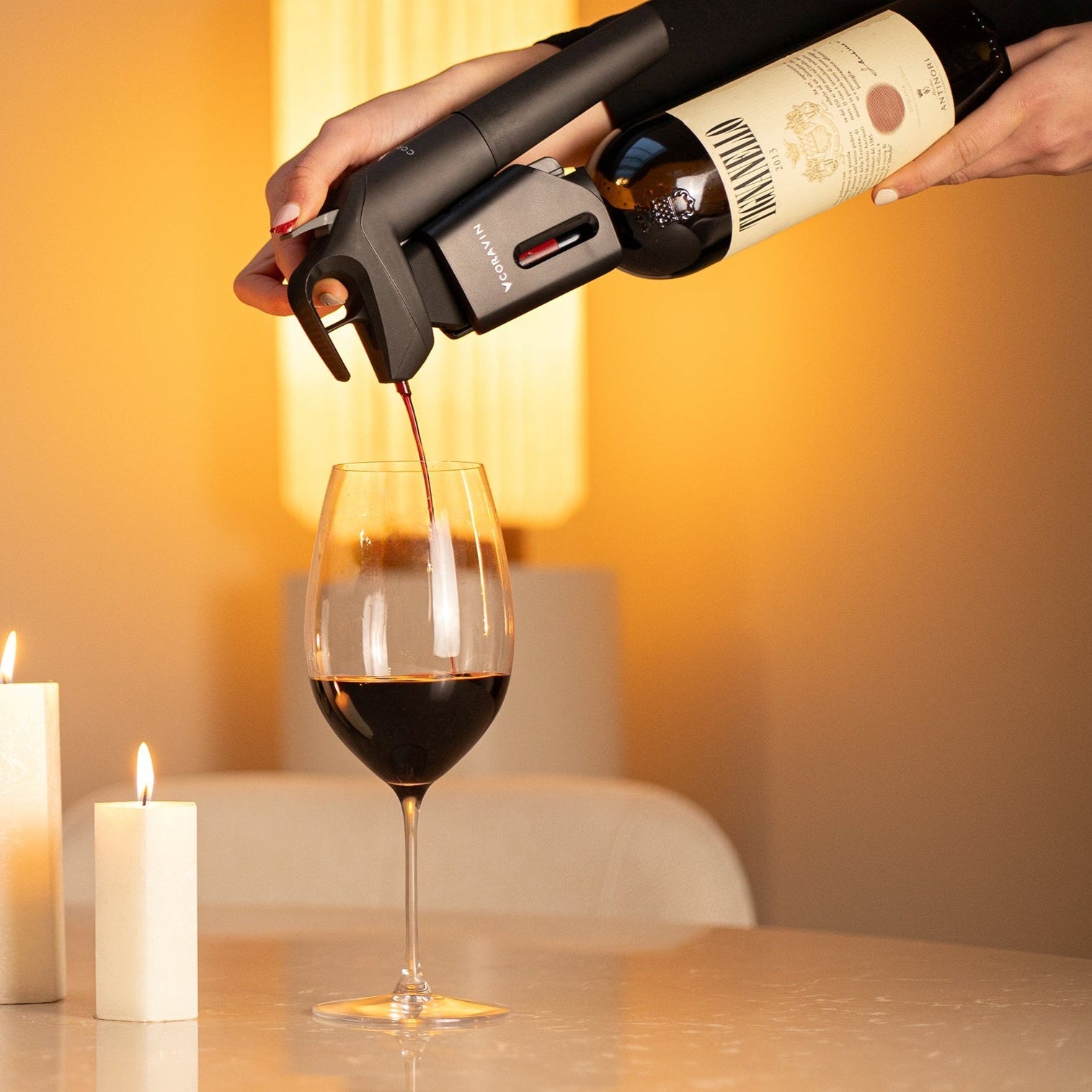 Person pouring red wine into a glass using a Coravin Timeless system, set on a marble table with lit candles and warm ambient lighting.
