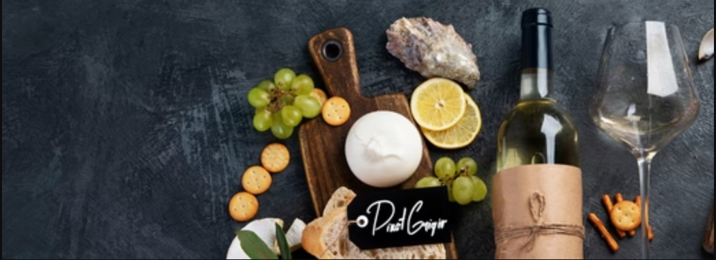 Flat lay of a Pinot Grigio wine pairing setup featuring a bottle of white wine, empty wine glass, green grapes, lemon slices, burrata cheese, crackers, bread, and an oyster on a dark textured surface.