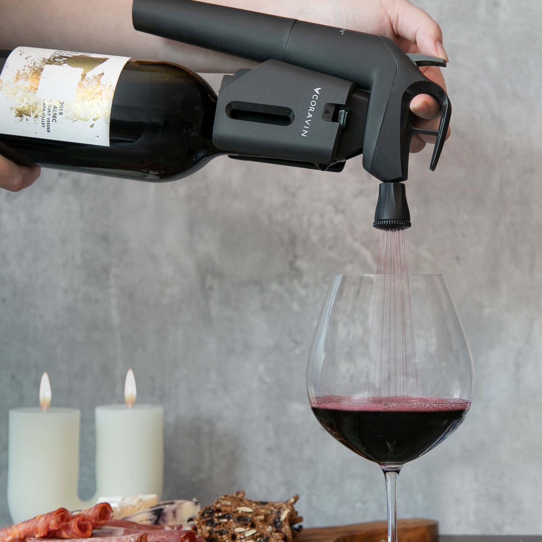 Red wine being poured into a glass using a Coravin Timeless system, with charcuterie board and candles in the background.