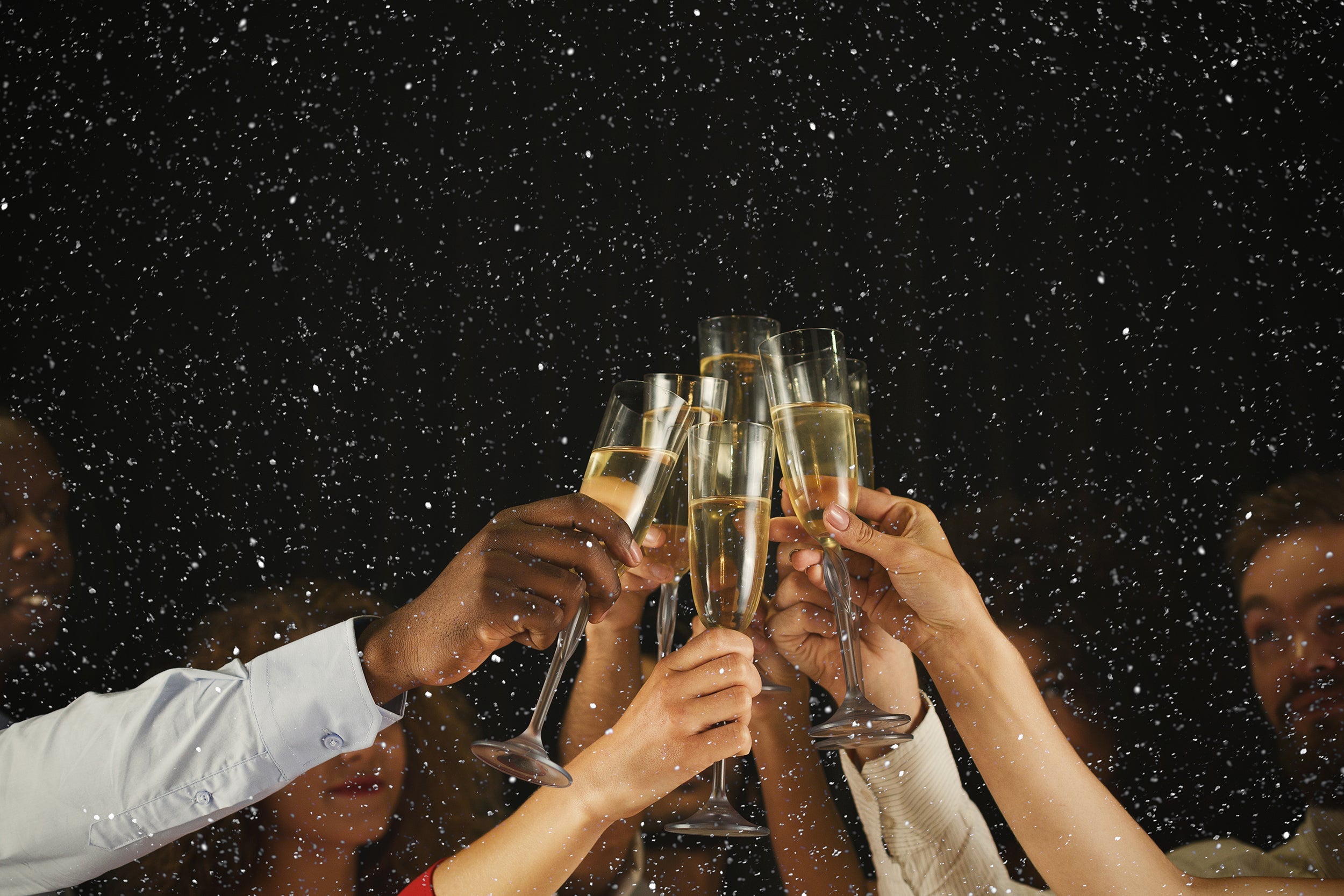 Group of people toasting with champagne flutes during a festive New Year celebration with snow falling against a dark background.