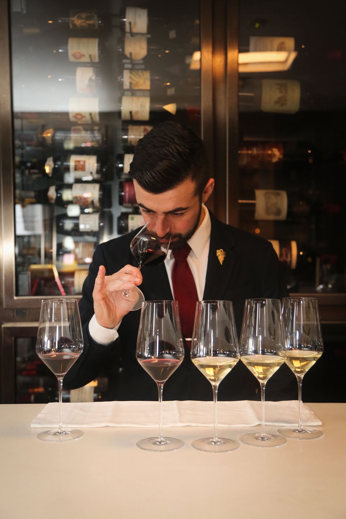 Sommelier in suit tasting red wine with a lineup of wine glasses in front of a wine cellar.