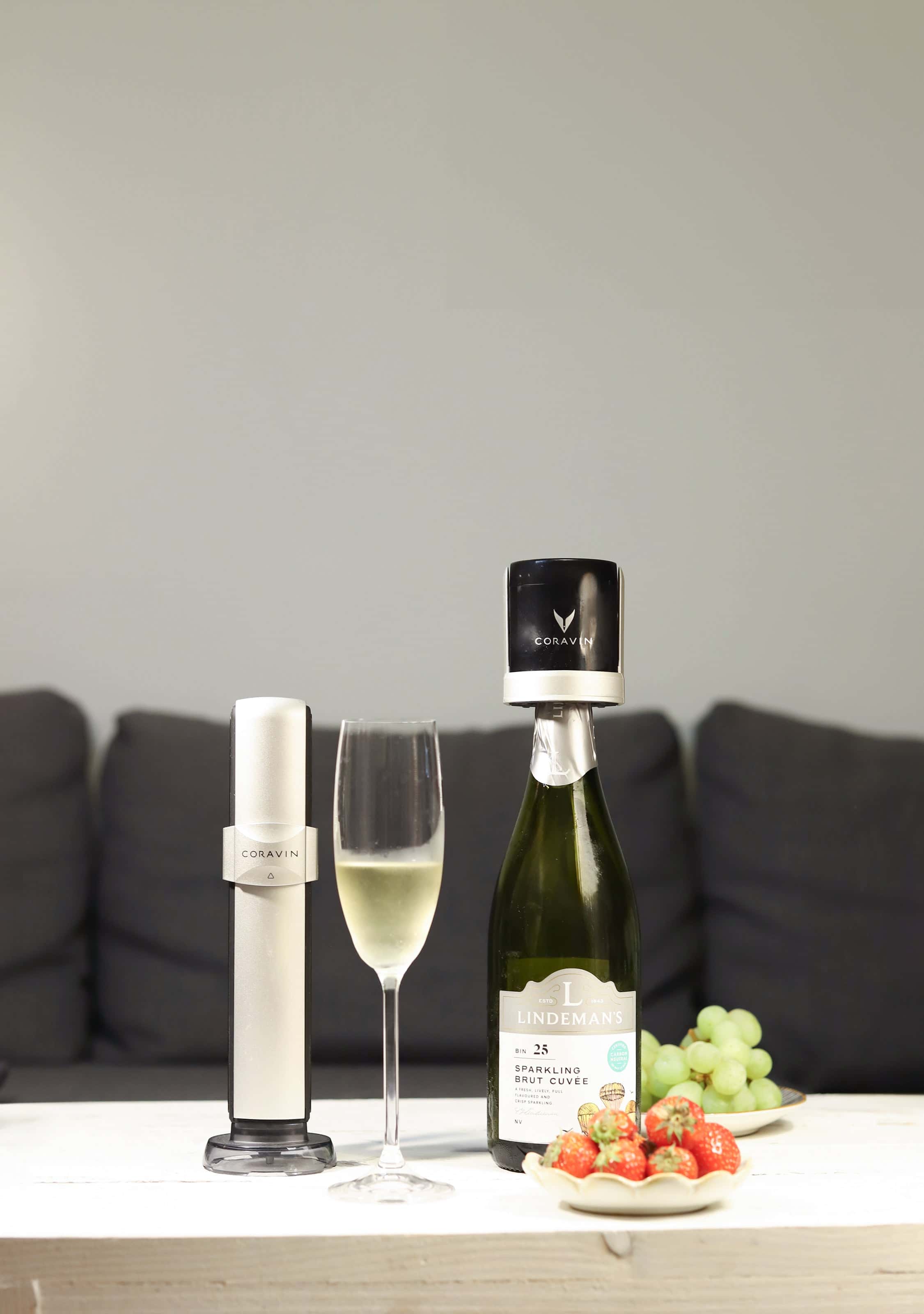 Coravin Sparkling wine preservation system displayed beside a bottle of Lindeman’s sparkling brut cuvée with a Coravin stopper, a filled champagne flute, and plates of fresh strawberries and green grapes on a white table in front of a dark sofa.