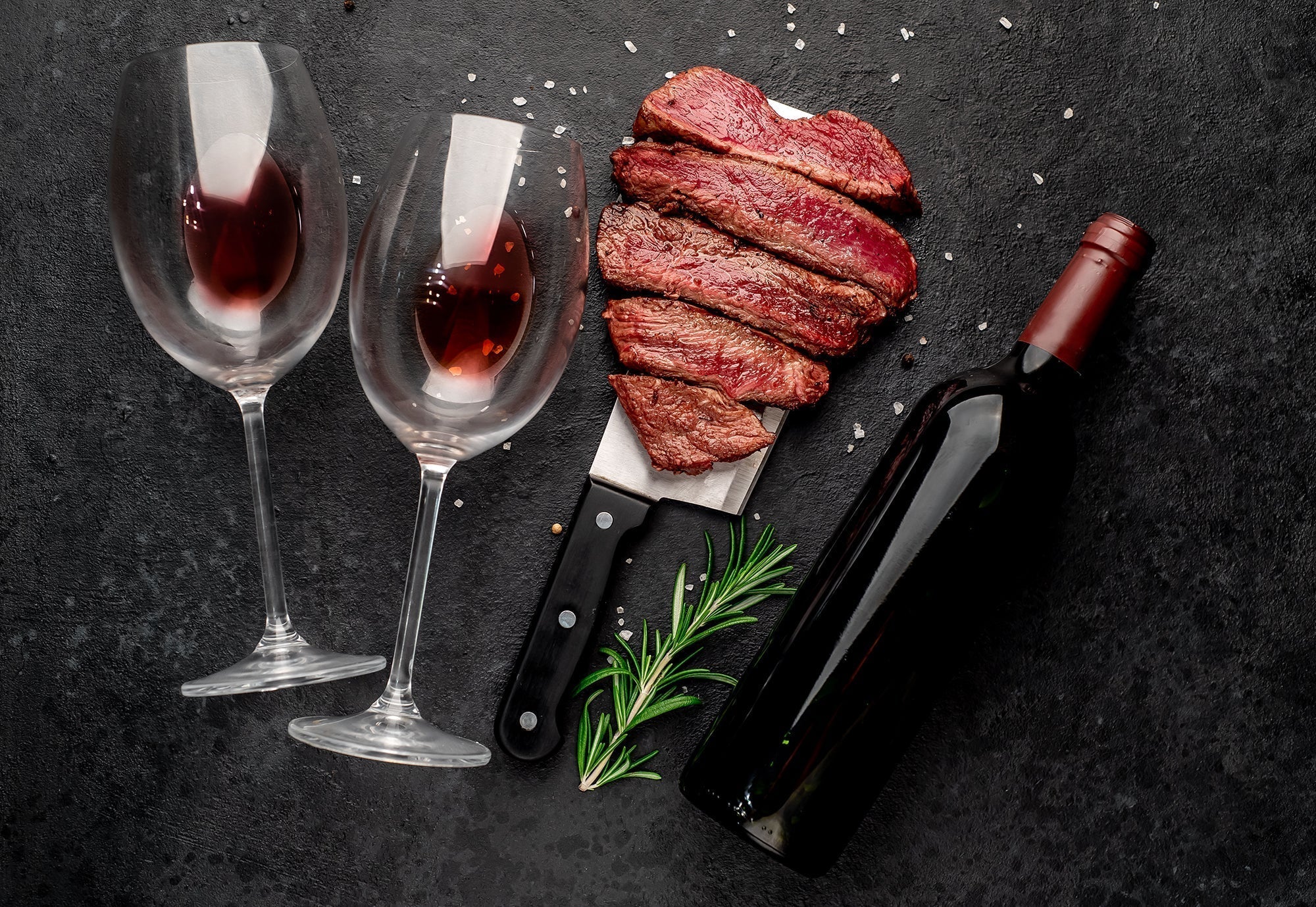 Romantic red wine and steak pairing with two glasses of red wine, sliced grilled steak shaped like a heart, rosemary garnish, and a bottle of wine on a dark stone background.