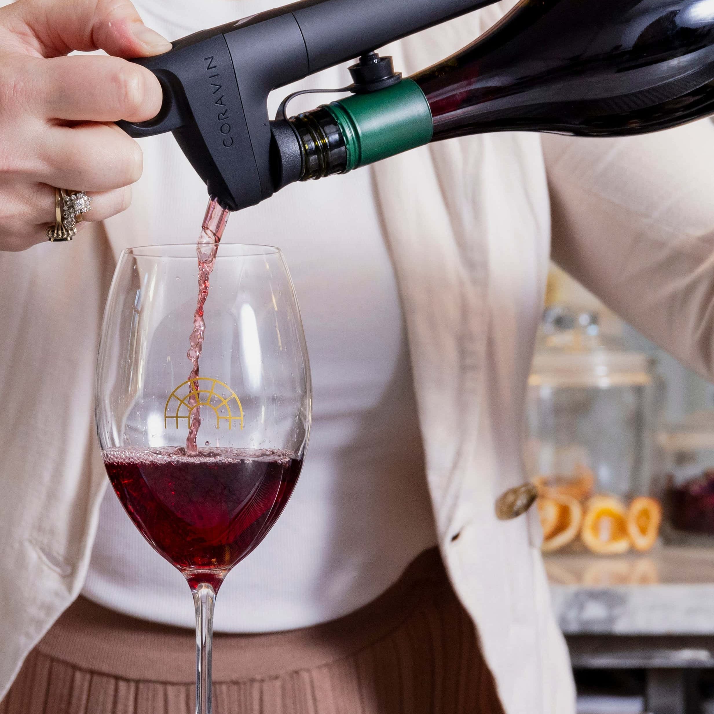 A woman pours red wine from a Coravin wine opener into a glass with a golden logo, set on a marble countertop.
