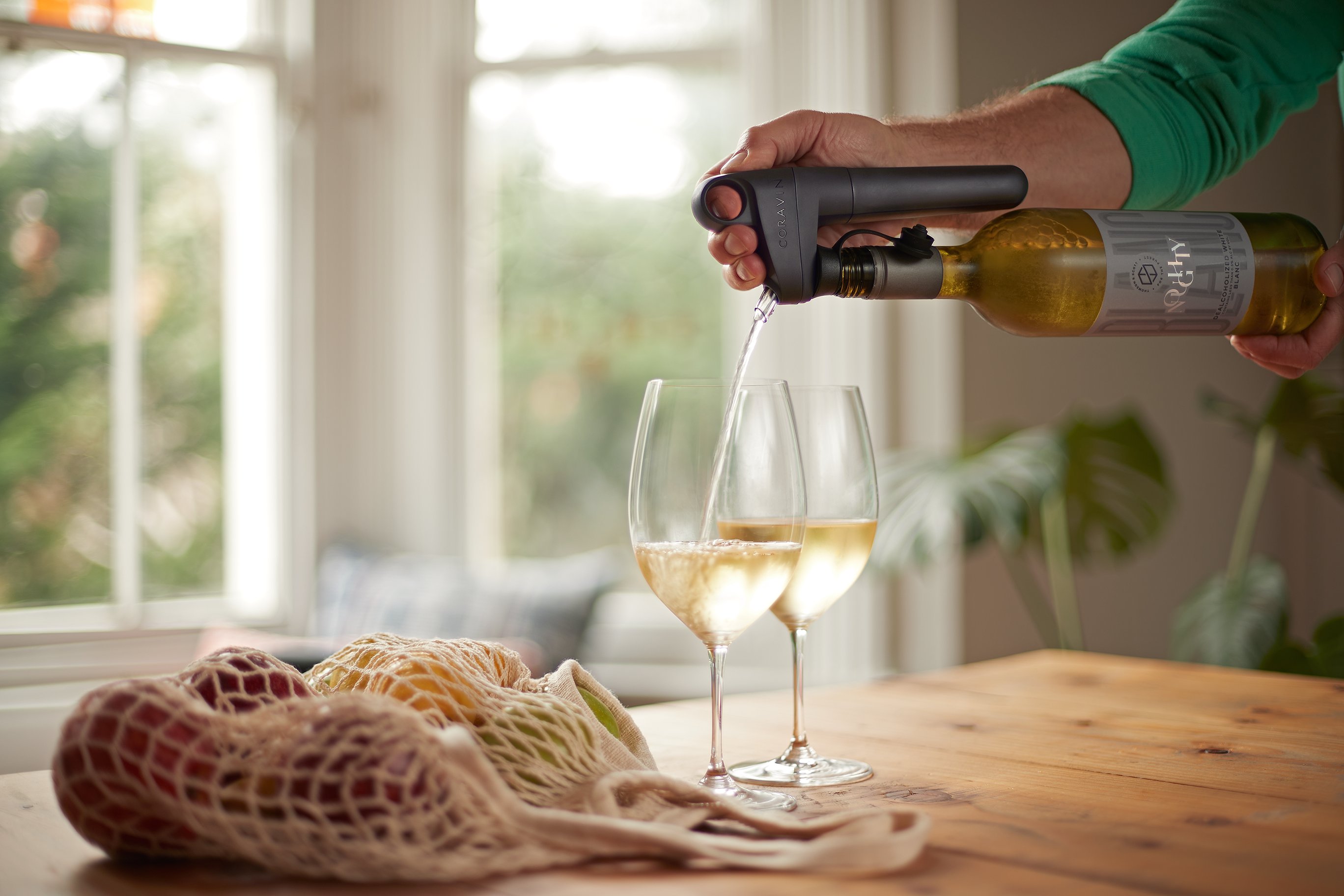 A hand pours white wine into two glasses using a wine preservation tool, with fruit in a mesh bag on a wooden table.